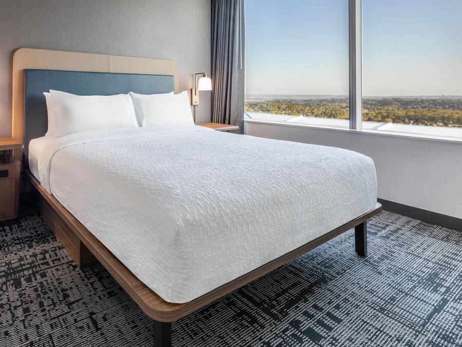 Habitación de hotel con una cama grande, una mesita de noche, una lámpara y vistas a un paisaje urbano a través de una ventana.