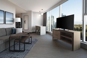 Ett modernt vardagsrum med en soffa, ett soffbord, en tv och ett skrivbord. Det finns stora fönster med utsikt över staden.
