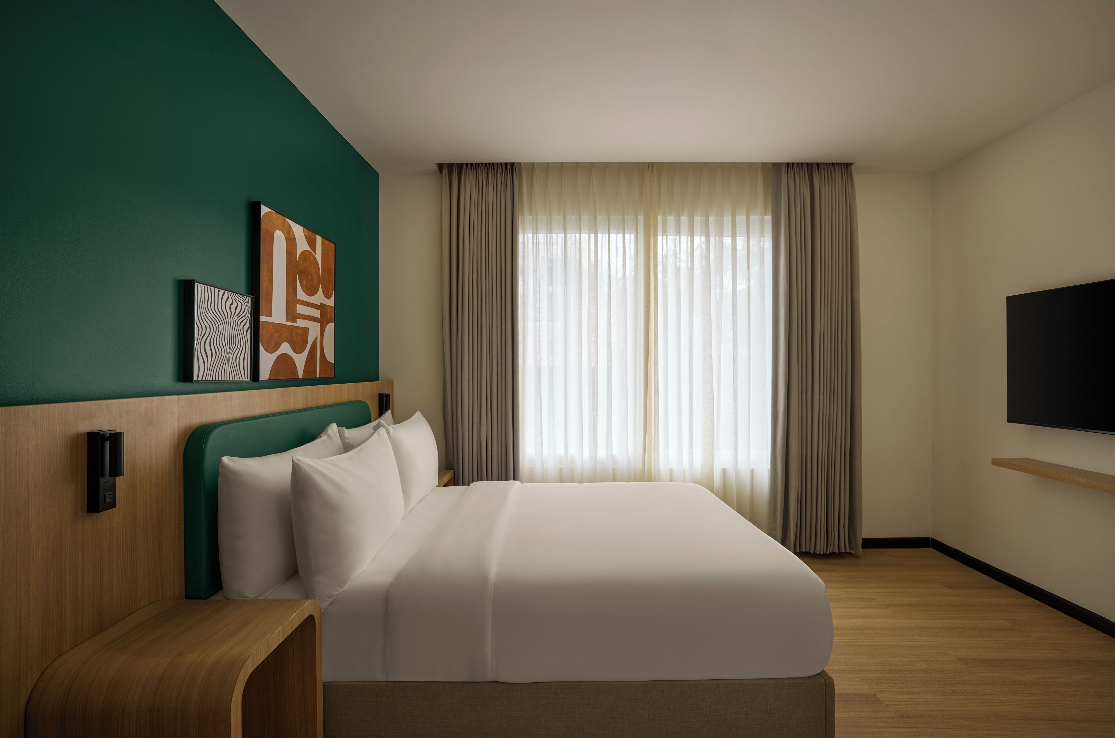 A hotel room with a bed, a TV, a wooden headboard, and artwork on the wall.
