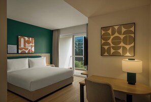 A hotel room with a bed, a desk, a chair, a lamp, and artwork on the walls.