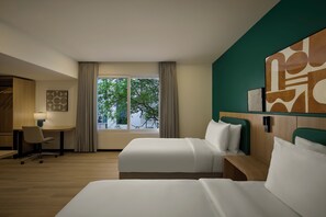 A hotel room with two beds, a desk, a chair, and a large window with a view of trees.
