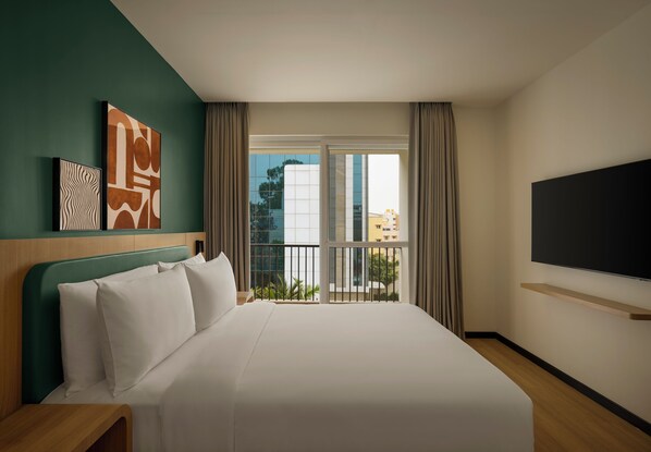 A hotel room with a large bed, a TV, artwork on the wall, and a view of buildings through the window.