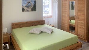Ein Zimmer mit einem Einzelbett, Holzrahmen, einer grünen Matratze, zwei Kissen, einem Nachttisch mit Lampe, einem weiteren Nachttisch mit Uhr, einem Holzschrank und einem Bild an der Wand.