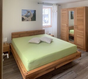 Habitació amb un llit individual de fusta, un matalàs verd, dos almoinos, una taula de nit amb una làmpada, una estant amb un rellotge, un armari de fusta i un quadre a la paret.