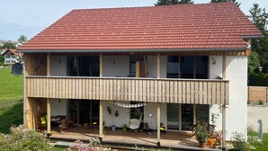 Ein zweistöckiges Haus mit rotem Ziegeldach, einem Balkon und einem Garten mit rosa Blumen.
