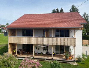 A two-story house with a red tiled roof, a balcony, and a garden with pink flowers. - Welcome to the apartment "Löwenzahn" in the house "Sonnige Auszeit" in Roßhaupten (Roßhaupten)