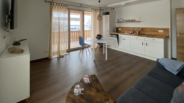 Ein Wohnzimmer mit grauem Sofa, einem Holztisch und einem Essbereich mit weißem Tisch und blauen Stühlen.