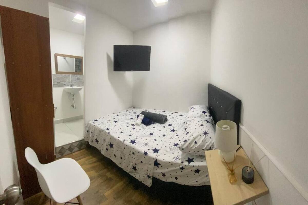 A small bedroom with a bed, a chair, a bedside table with a lamp, a TV on the wall, and a bathroom visible through an open door.