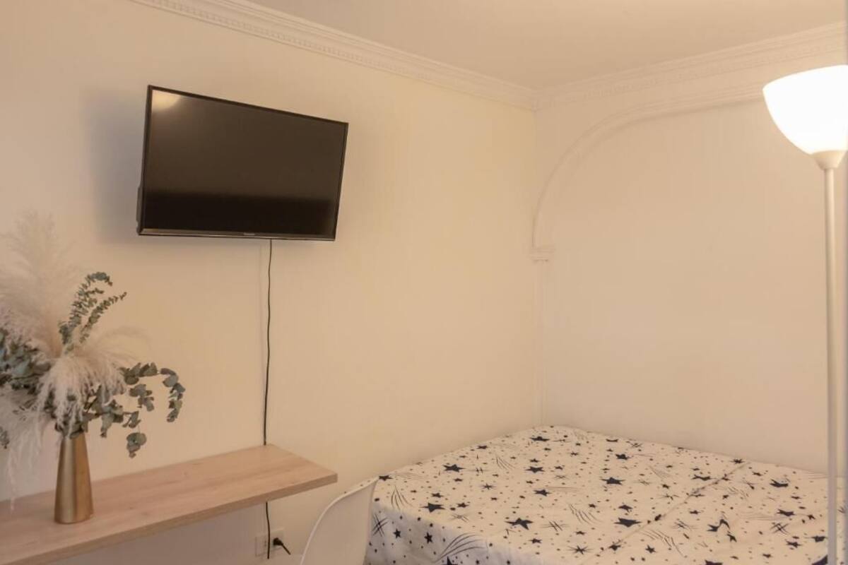 A room with a bed, a TV mounted on the wall, a bedside table with a vase, and a lamp.
