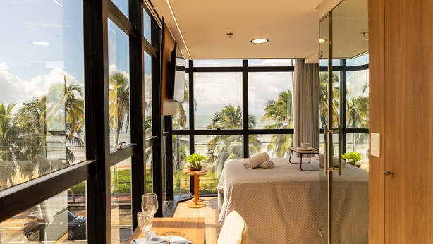 A modern hotel room with large windows, a bed, a small table with a plant, and a view of palm trees. - Setai Edition #206 - Waterfront by Carpediem (João Pessoa)