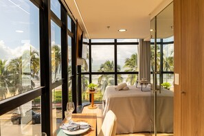 Une chambre d’hôtel moderne avec de grandes fenêtres, un lit, une petite table avec une plante et une vue sur des palmiers.