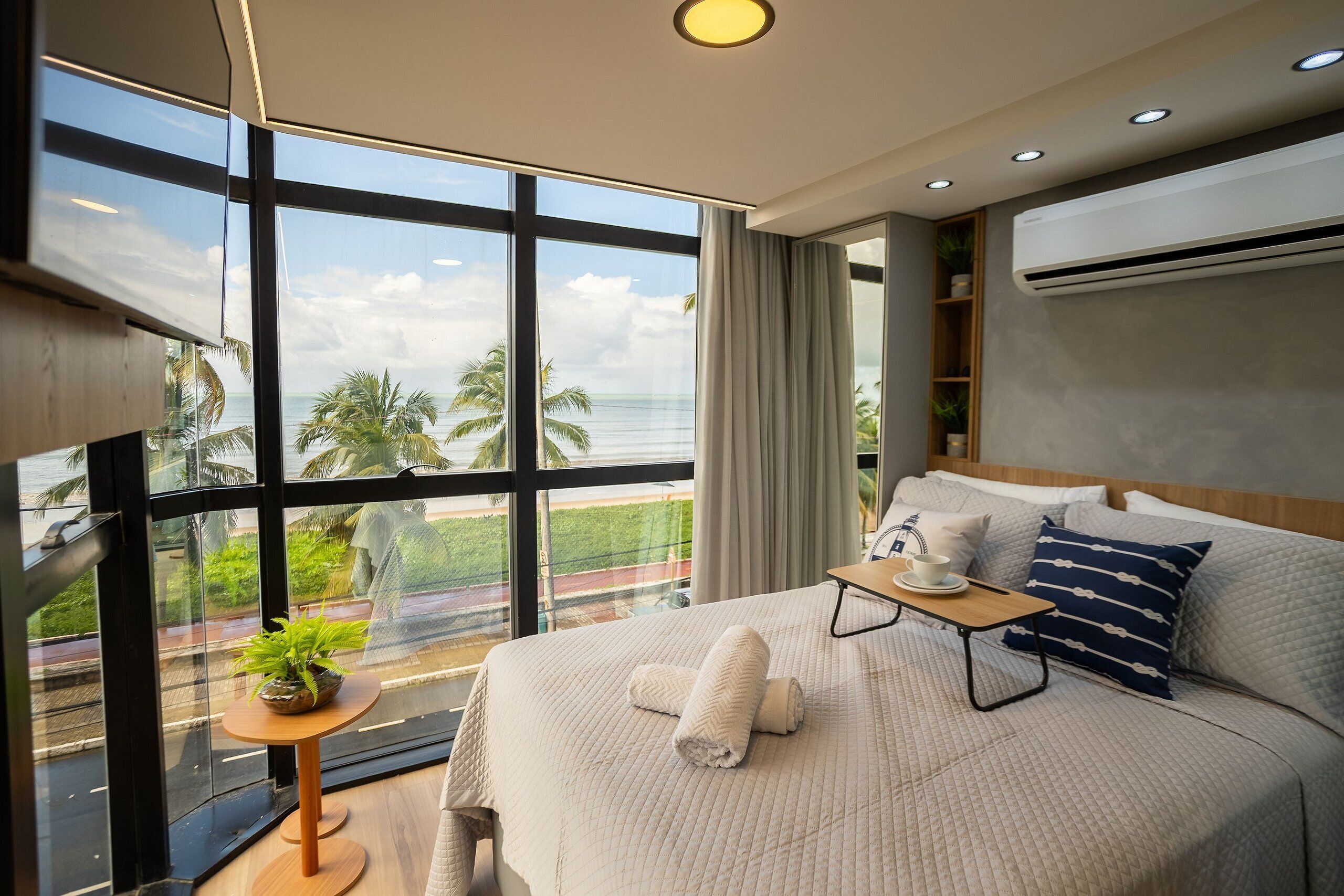 Une chambre d’hôtel moderne avec une grande fenêtre, un lit recouvert d’une couette blanche, une table de chevet agrémentée d’une plante et un appareil de climatisation.