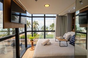 Ein modernes Zimmer mit großem Fenster, einem Bett mit Kissen, einem kleinen Tisch mit Tasse und einem an der Wand befestigten Fernseher.