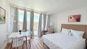 A hotel room with a bed, a dining table with chairs, a small table with a plant, and a view of the city through large windows. - Cozy Studio at St. Johns – Steps to Beach (Myrtle Beach)