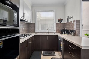 A compact kitchen with dark wooden cabinets, a stainless steel sink, and a window with blinds. - Spacious 3-bed Townhouse in Coburg (Melbourne)