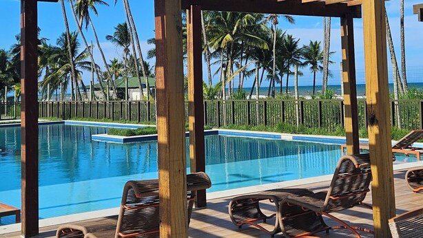 A poolside area with wooden pergolas, lounge chairs, and a clear view of palm trees and a pool. - Apê Maré Blue - Seu Paraíso à beira-mar! (ILHEUS)