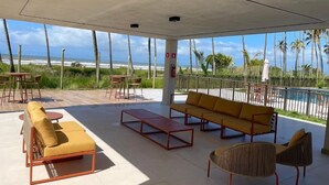 A covered outdoor seating area with a pool view, including a sofa, chairs, and a table. - Apê Maré Blue - Seu Paraíso à beira-mar! (ILHEUS)
