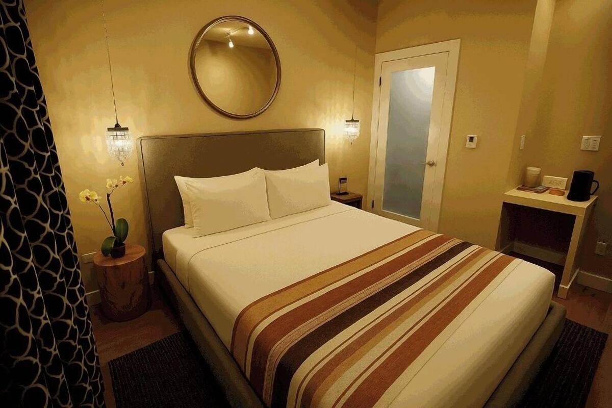 A hotel room with a bed, a round mirror, a nightstand, a small table with a lamp, and a door.