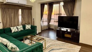 Uma sala de estar com um sofá verde, uma TV de tela plana, um candelabro e uma grande janela com cortinas.