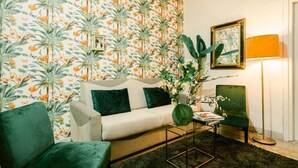 Ein Wohnzimmer mit blumigem Wandpapier, einer Couch, einem grünen Sessel, einem Couchtisch und einer Stehlampe.