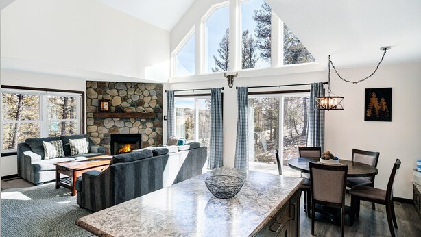 A spacious living area with a stone fireplace, a large window, and a dining area with a marble countertop. - Cozy Cabin: Hot Tub + Fireplace, Near Breck (Fairplay)