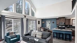 A spacious living area with a sofa and a coffee table, a kitchen with a bar area, and large windows offering a view of trees. - Cozy Cabin: Hot Tub + Fireplace, Near Breck (Fairplay)