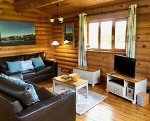Ein Holzblockhaus-Innenraum mit einer Ledersofa, einem Fernseher auf einem Holzbodenständchen, einer Lampe und einem Gemälde an der Wand.