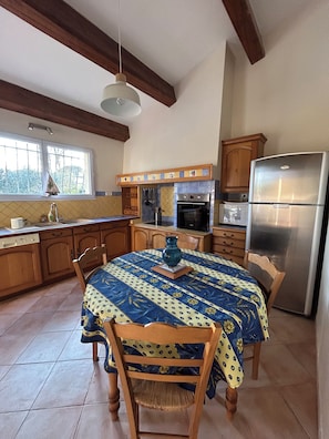 A kitchen with wooden cabinets, a stainless steel refrigerator, a dining table with a patterned tablecloth, and a hanging light fixture.