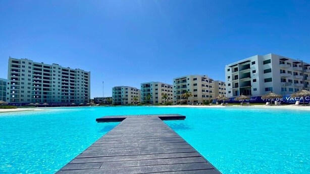 A wooden deck leading into a clear blue pool with a beach and resort buildings in the background. - Apartment with balcony overlooking crystal-clear lagoon and (Mazatlán)