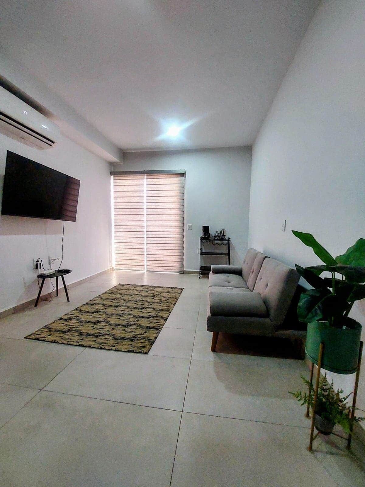 Una sala de estar con sofá, televisor montado en la pared, ventana con persianas, una mesita y una planta en maceta.