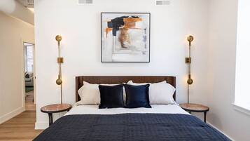 A modern bedroom with a large bed, a headboard, two bedside tables, a wall-mounted abstract painting, and a window.