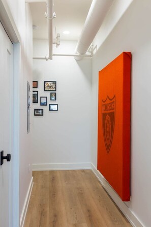 Un couloir avec un plancher en bois, des murs blancs et une porte sur la gauche. Des tableaux encadrés ornent le mur, ainsi qu’une grande bannière orange portant un logo en forme d’écusson.