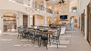 A spacious dining area with a large wooden table and black chairs, a stone fireplace, and a high ceiling with exposed beams.
