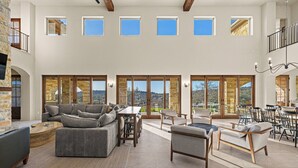 A spacious living area with a gray sectional sofa, a wooden coffee table, and a dining area with a table and chairs. Large windows provide views of the outdoors.