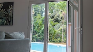 Una stanza con un divano grigio, una porta vetrata scorrevole che dà su una piscina e vista su un giardino tropicale.
