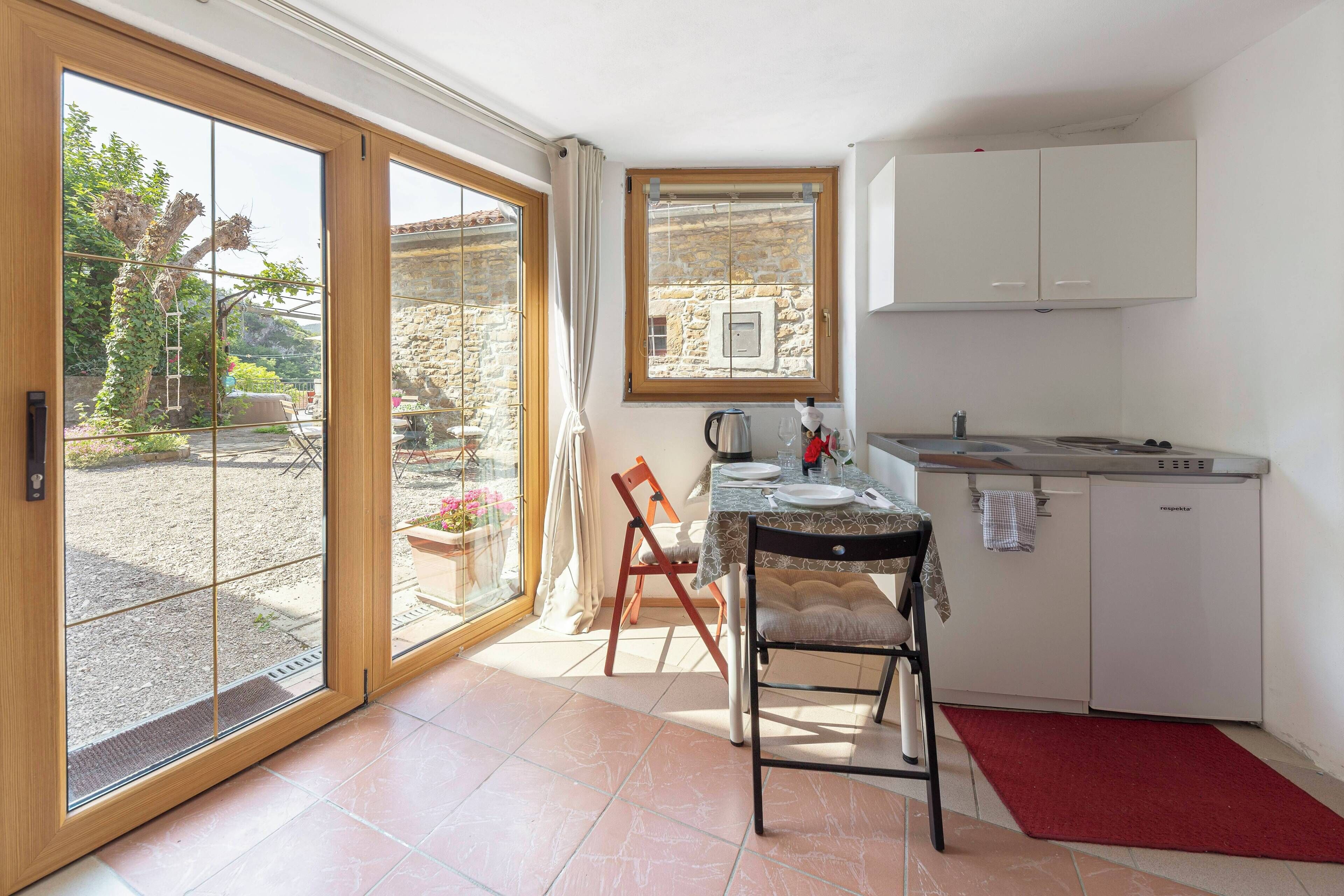 A compact kitchen with a dining area, a window with a view of a garden, and a small refrigerator.