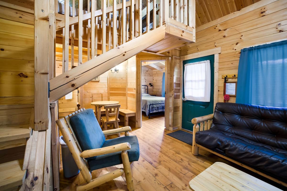 A wooden cabin interior with a staircase, a sofa, a dining table, and a bed.