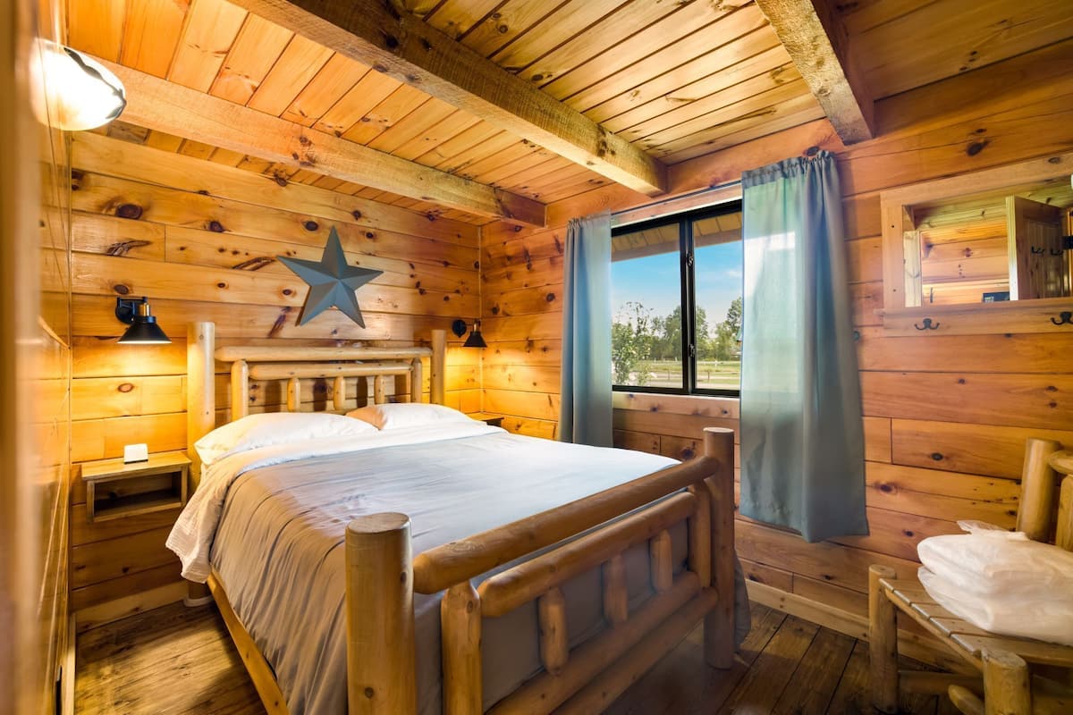 A wooden cabin bedroom with a bed, bedside lamps, a star decoration, a window with curtains, and a view of the outdoors.
