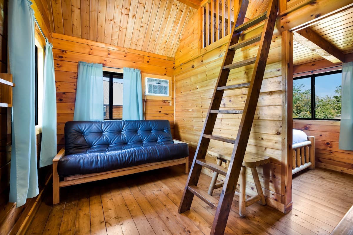 A wooden cabin interior with a sofa, a ladder, and a window with curtains.
