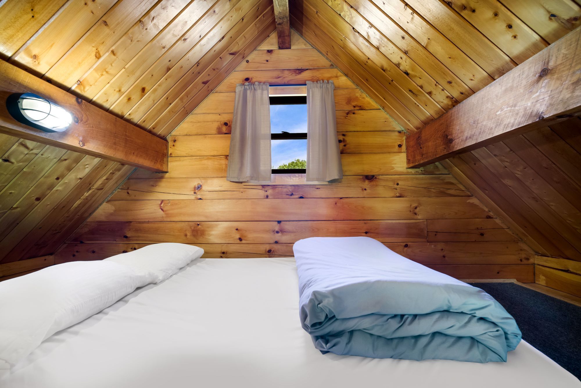 A cozy attic bedroom with a wooden sloped ceiling, a single bed with white and blue bedding, and a small window with curtains.