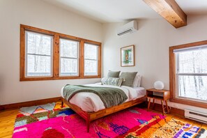 A bedroom with a wooden bed, colorful rug, large windows, and a small bedside table.