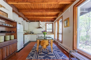 A kitchen with wooden beams, a dining table with chairs, and a view through large windows.