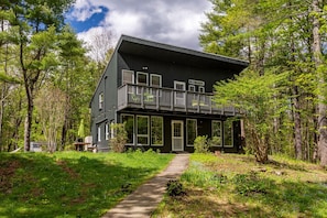 A two-story house with a balcony, surrounded by a forested area.