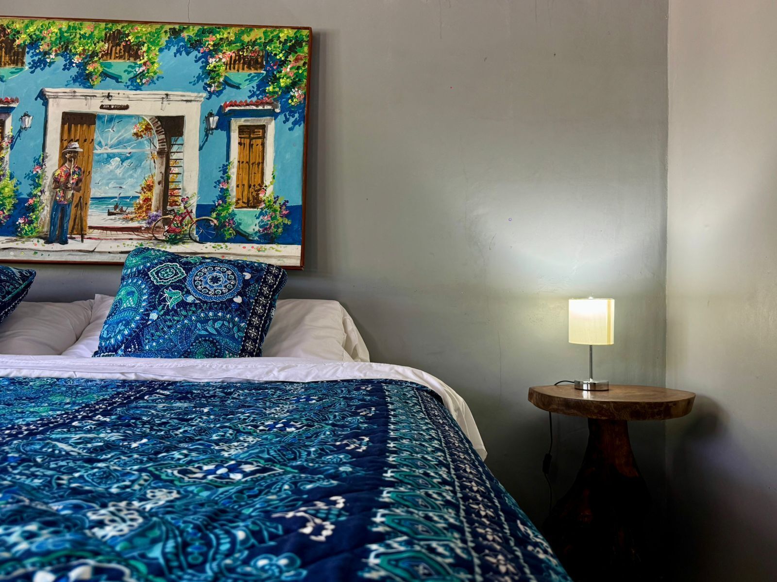 Una camera da letto con un letto, cuscini, un comodino, una lampada e un quadro appeso al muro.