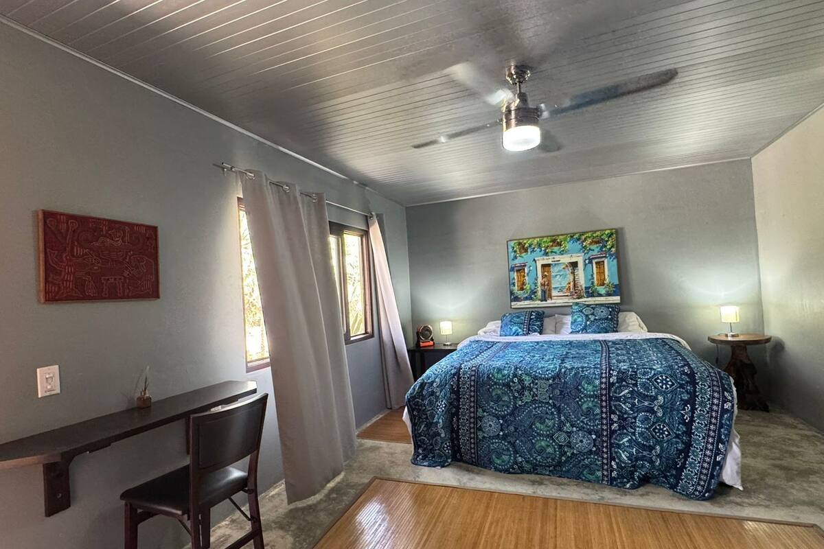A bedroom with a bed, a desk, a chair, a ceiling fan, and a painting on the wall.