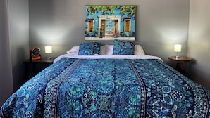 Quarto com uma cama grande, uma pintura de um edifício, duas luminárias de cabeceira e uma pequena mesa com um ventilador.
