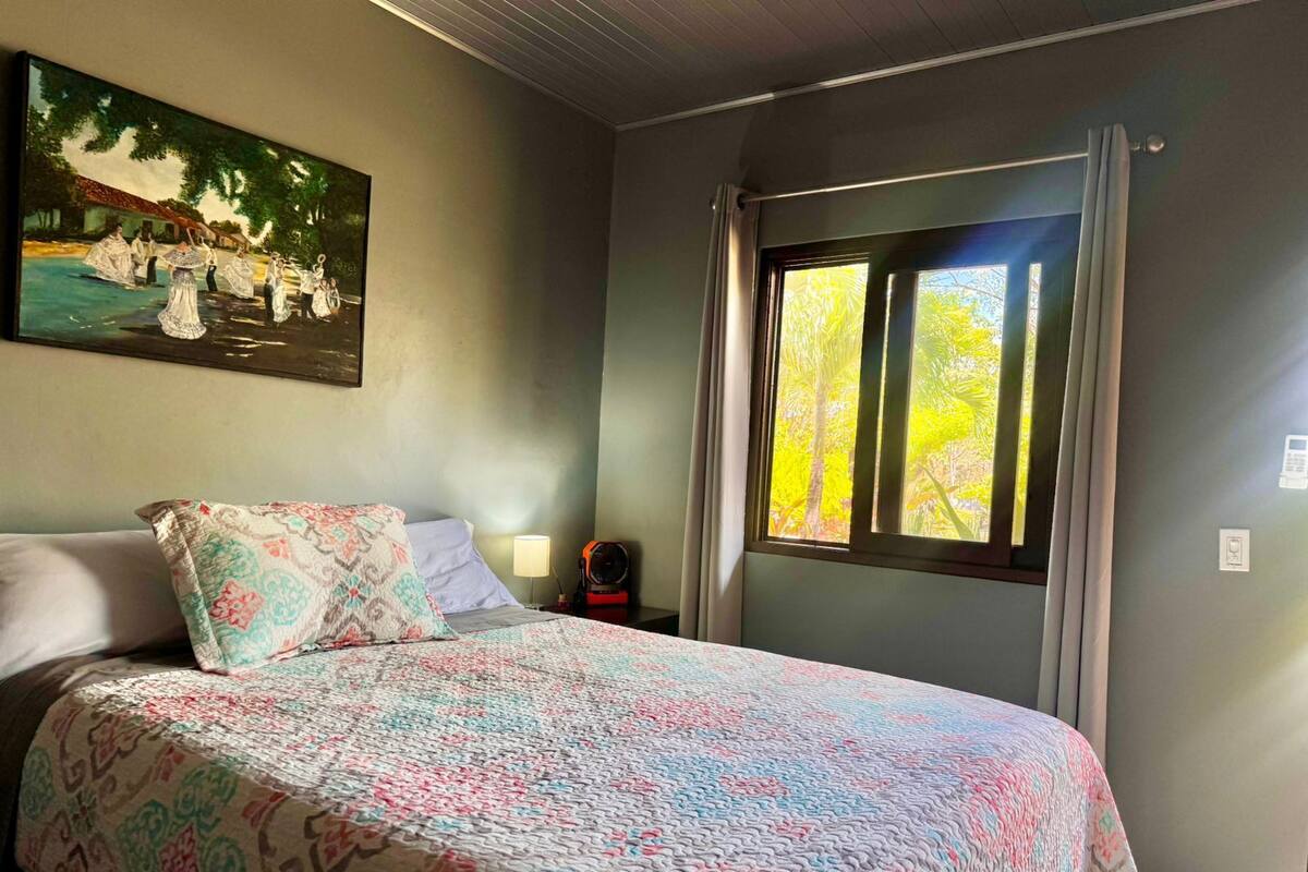 A bedroom with a bed, a painting on the wall, a window with curtains, and a nightstand with a lamp.