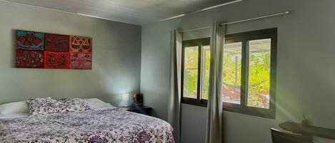 A bedroom with a bed, a desk, a chair, a ceiling fan, a window with curtains, and a wall with artwork.