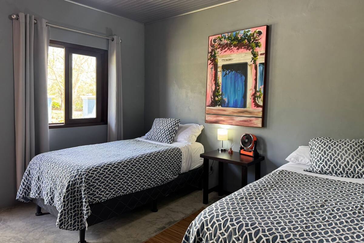 A room with two beds, a window with curtains, a table with a lamp and fan, and a painting on the wall.