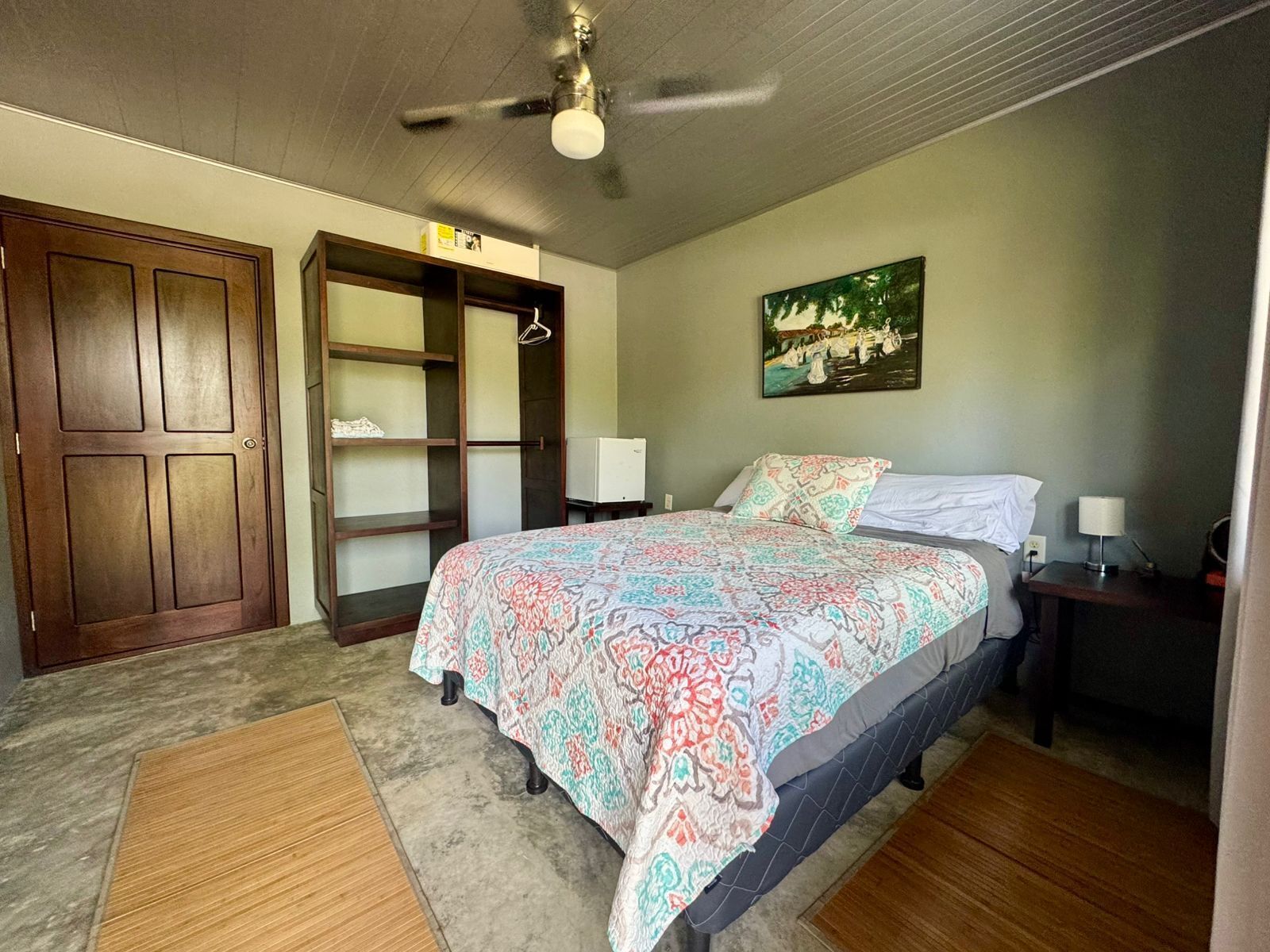 Un dormitorio con cama, ventilador de techo, armario de madera y un cuadro en la pared.